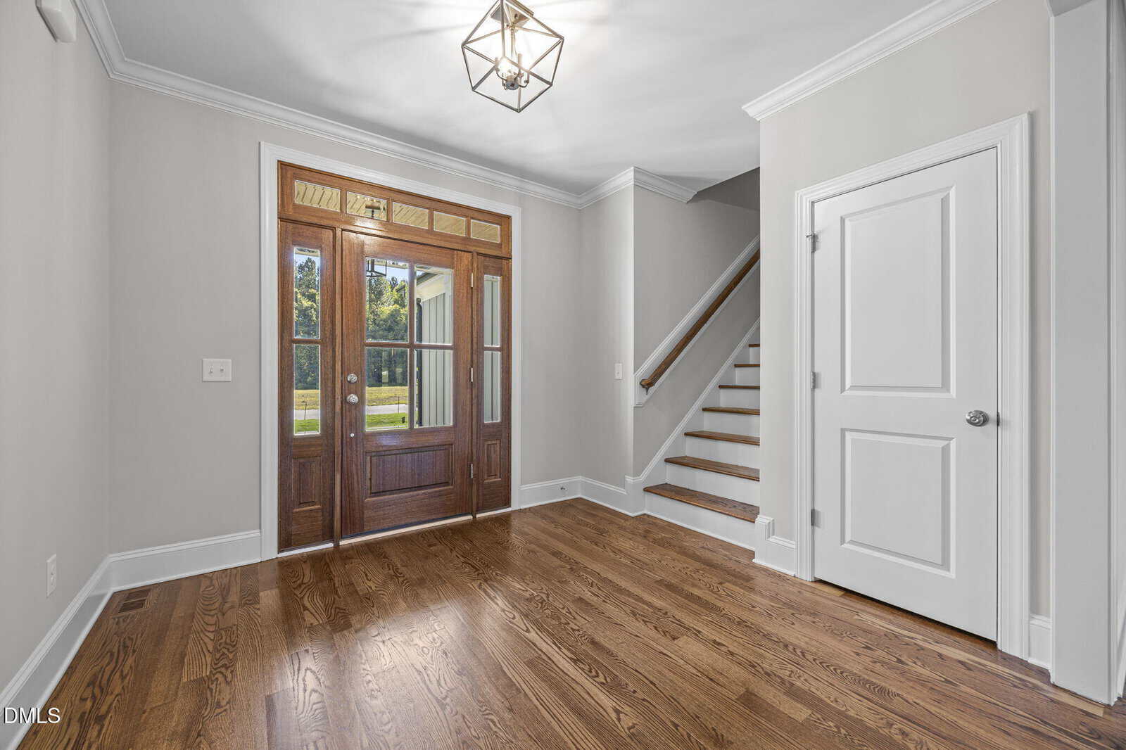 A foyer with hardwood floors, a wooden front door with glass panels, white walls, a white door to the right, and a staircase leading upstairs. A geometric light fixture hangs from the ceiling.