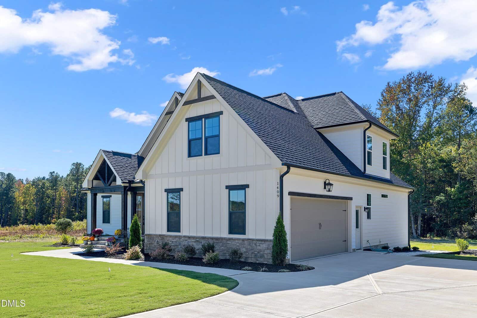A modern two-story house with light-colored siding, dark roof shingles, black trim, and a two-car garage. The home sits on a manicured lawn with a concrete driveway and small landscaped garden beds.
