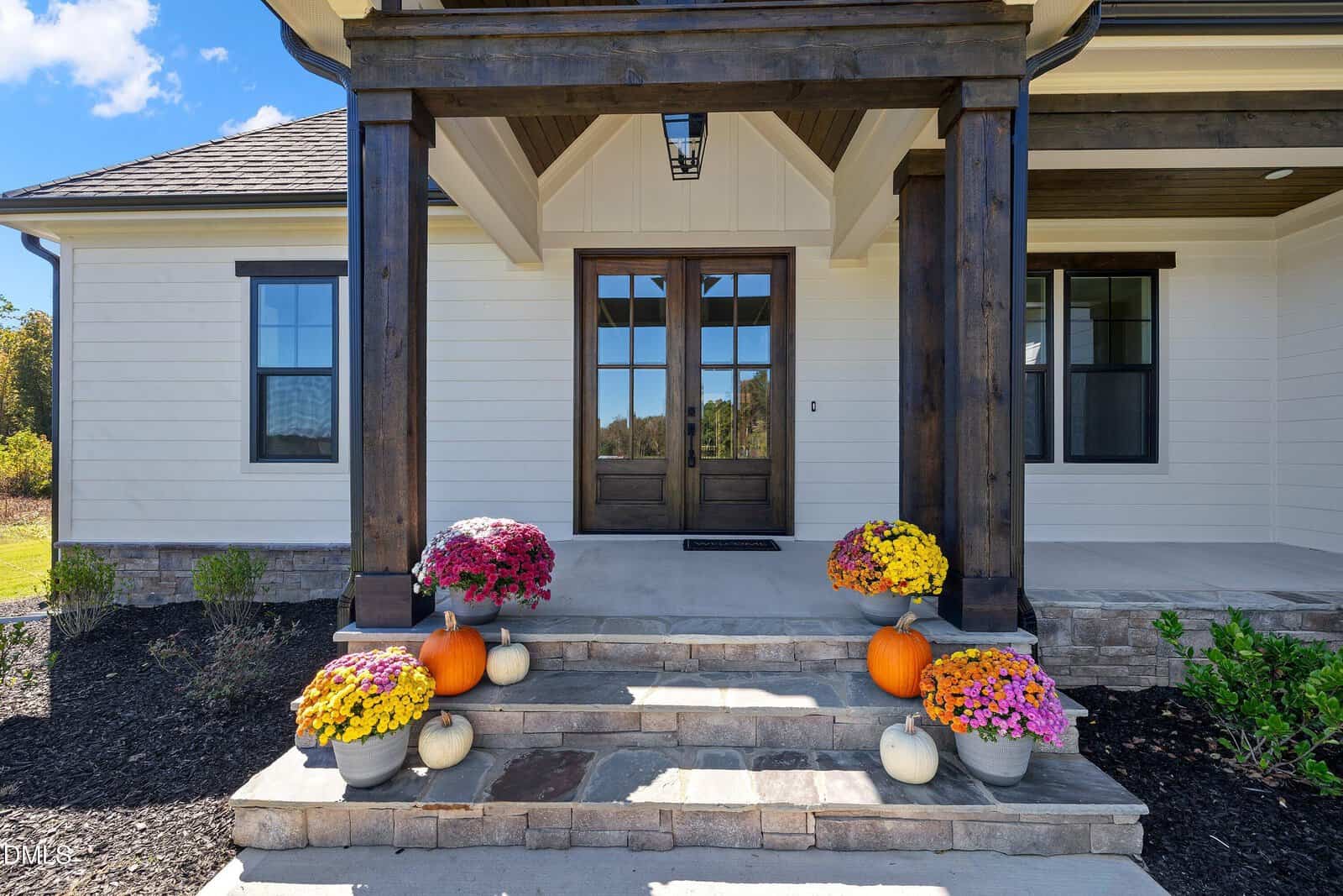 The front porch of a house with wooden double doors, stone steps, and potted yellow, purple, and pink flowers. Orange and white pumpkins are arranged on both sides of the steps.