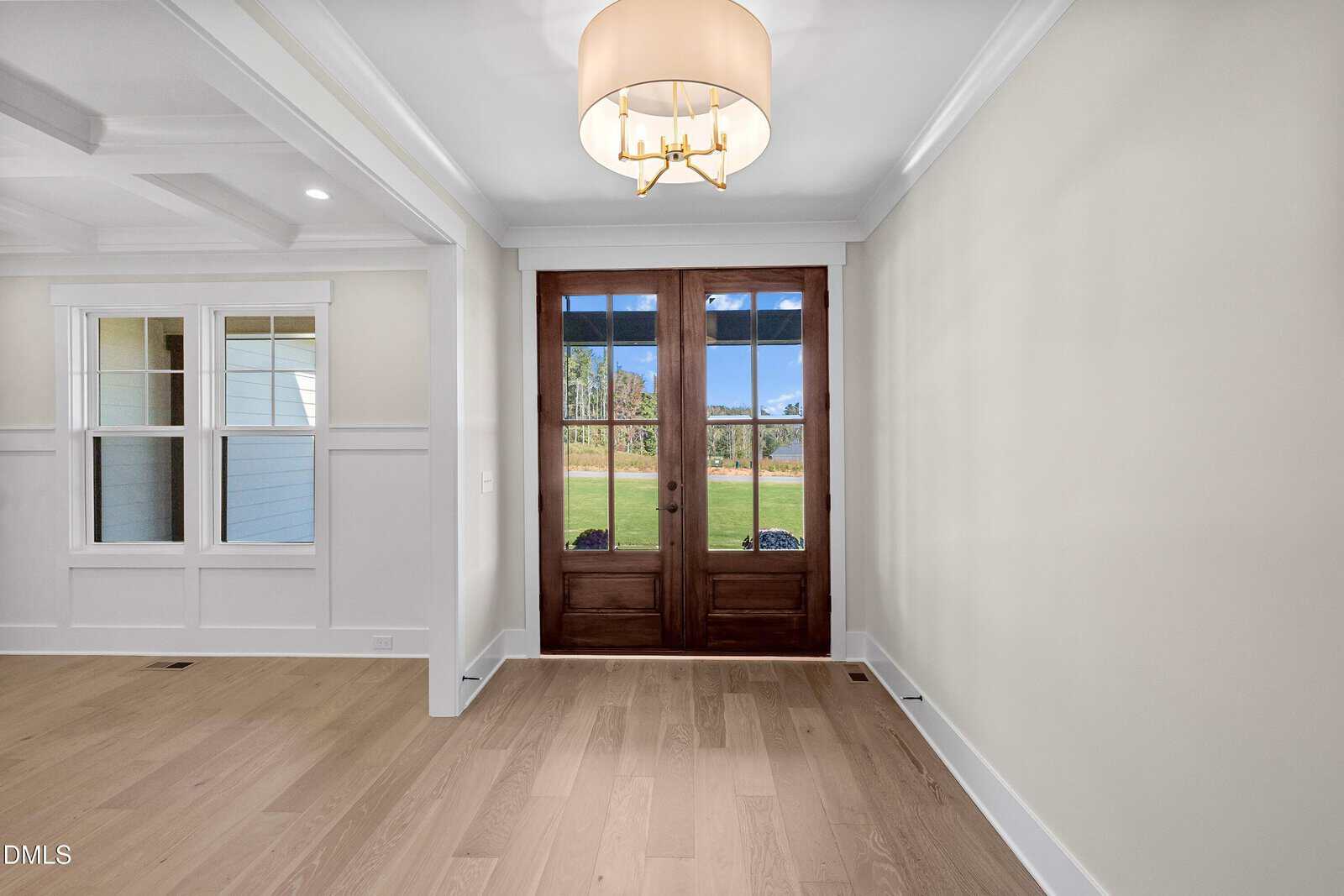 A room with light wood floors, cream-colored walls, a coffered ceiling, and a modern chandelier. Double glass doors with dark wood frames open to a grassy outdoor area. Adjacent are three interior windows.