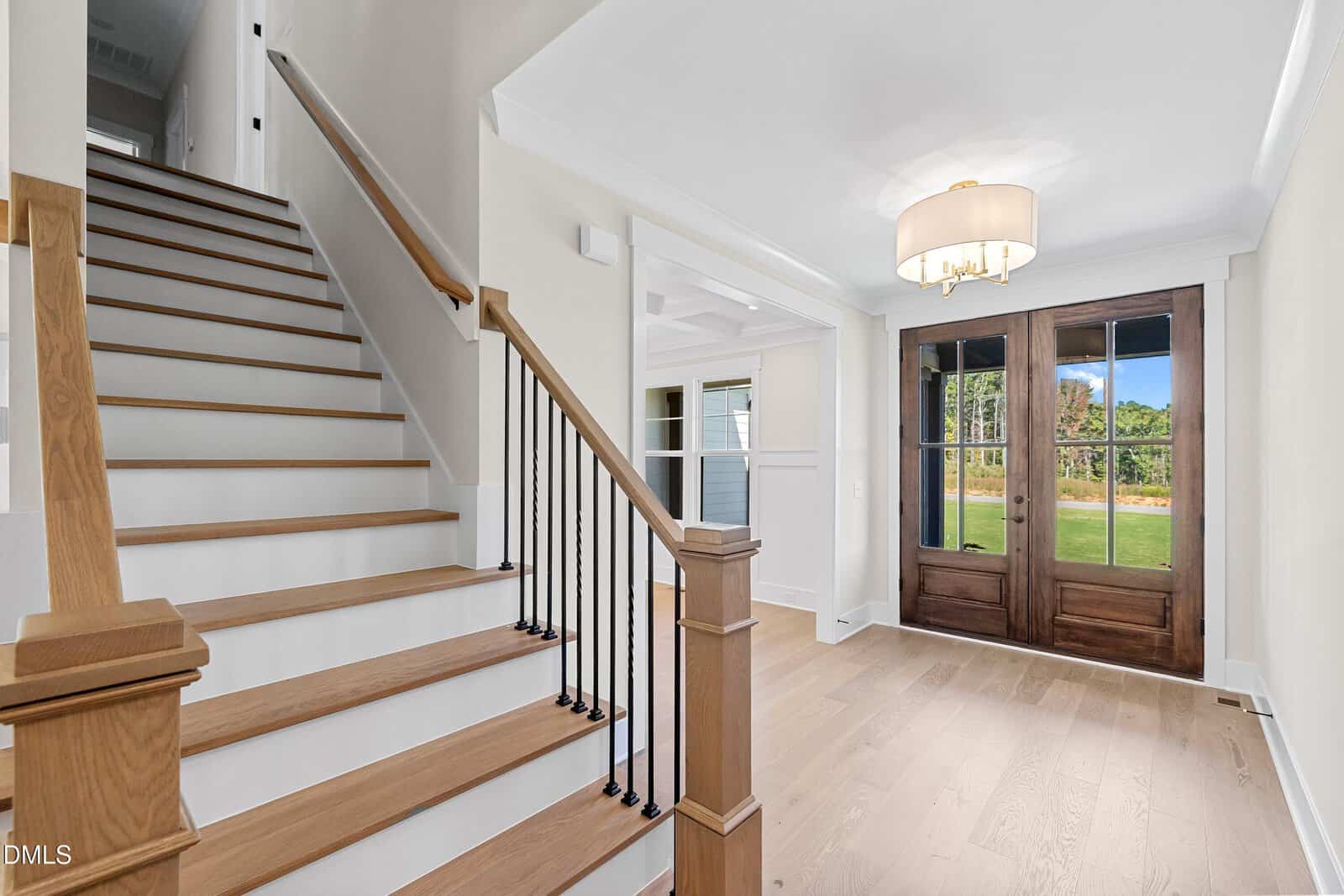 A bright entryway with light wood floors, a wooden staircase with black balusters, white walls, a large light fixture, and double front doors with glass panels, looking out to a green lawn.