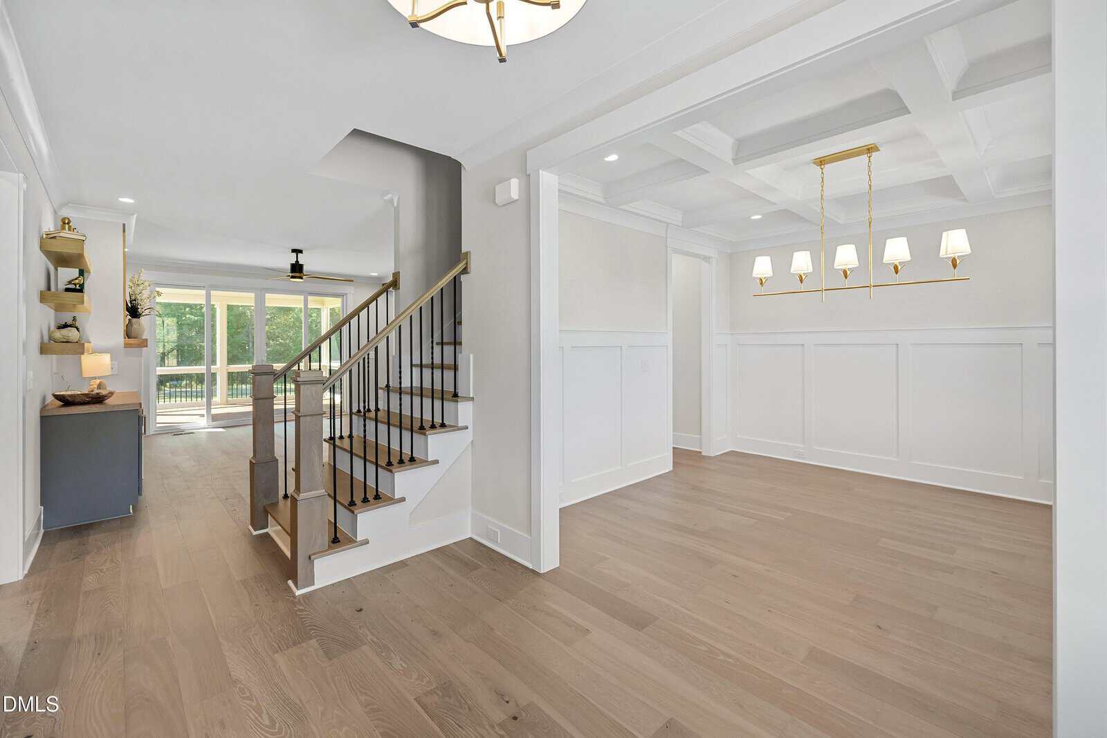 A bright, open interior features light wood floors, a staircase with dark railings, a dining area with a coffered ceiling and chandelier, and large windows leading to a patio with a view of greenery.