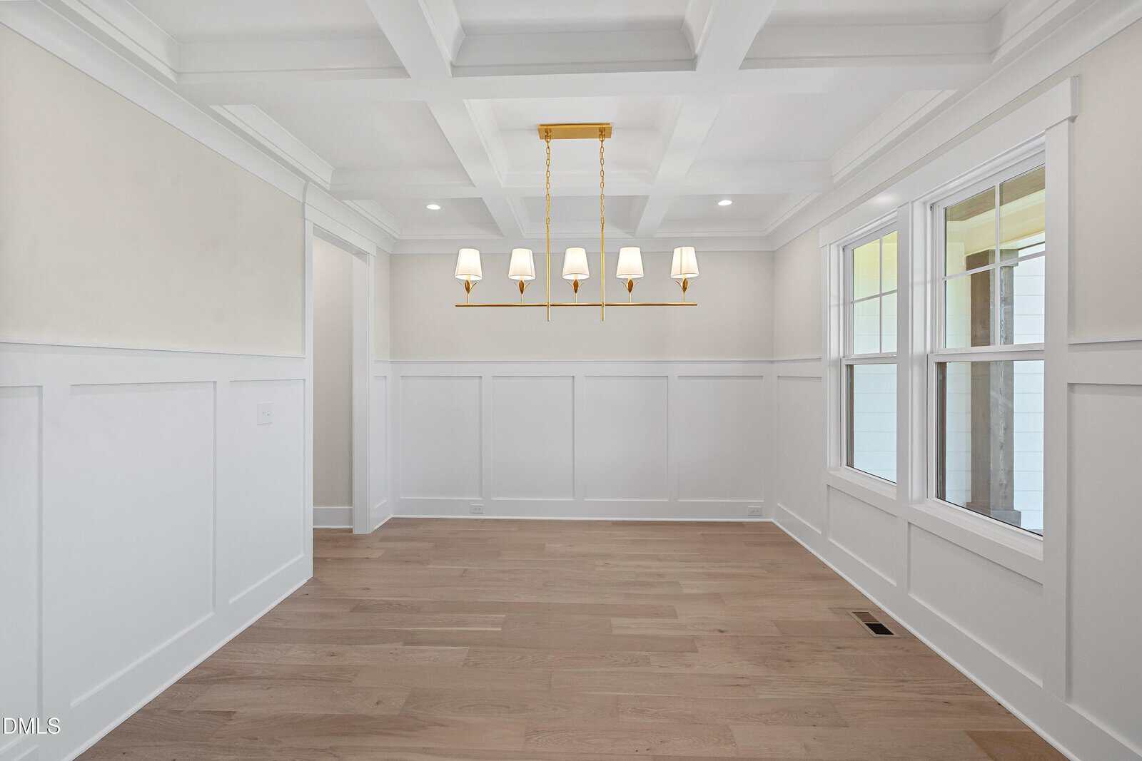 A bright, empty dining room with white wainscoting, a coffered ceiling, light wood flooring, large windows, and a gold chandelier with six lampshades hanging in the center.