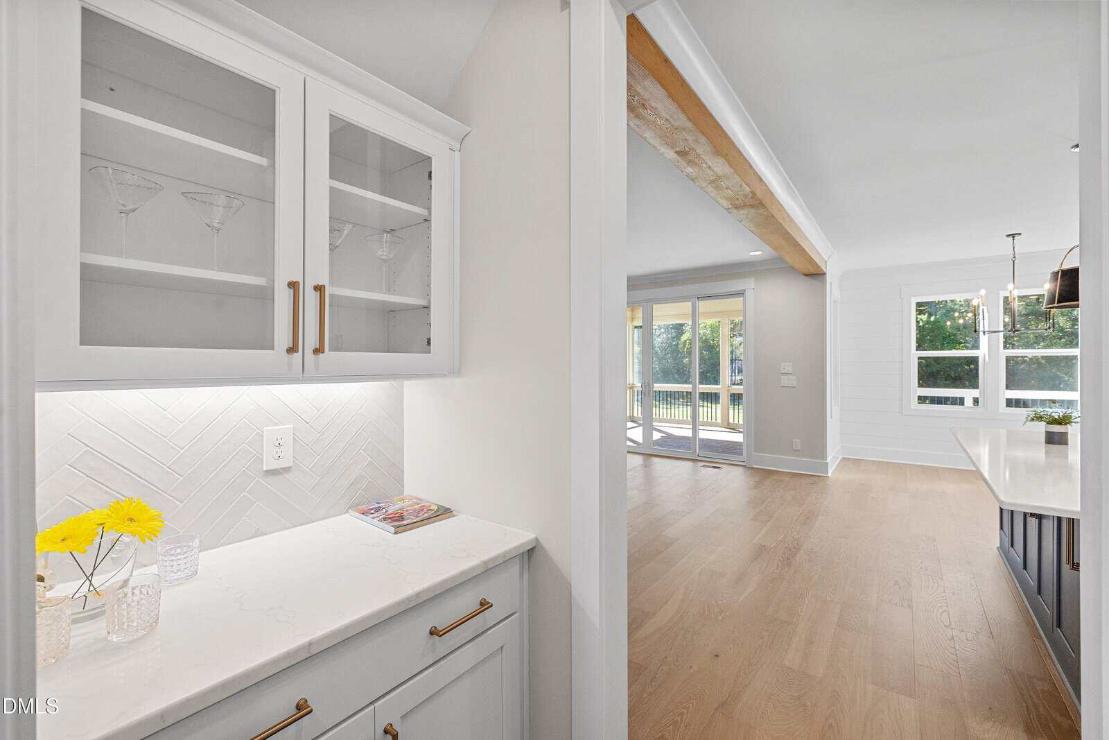 Bright kitchen nook with white cabinets, glass doors, and gold handles. Marble countertop holds a vase with yellow flowers and magazines. Open doorway leads to a spacious living area with large windows and light wood floors.