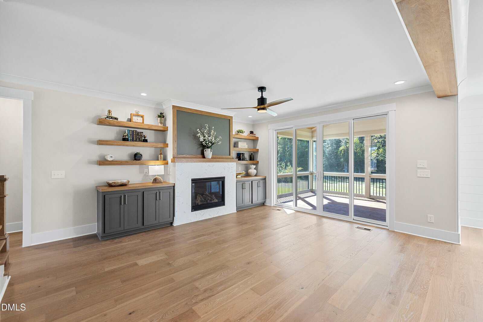 A modern living room with light wood floors, built-in shelves and cabinets flanking a white fireplace, a ceiling fan, and large sliding glass doors opening to a backyard with trees.