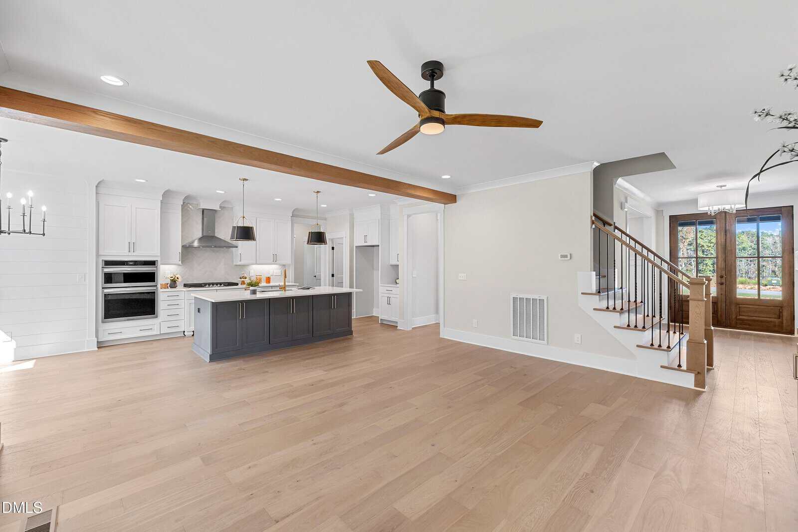 Open-concept kitchen and living area with light wood floors, white cabinets, dark island, modern ceiling fan, wooden ceiling beam, and stairs leading to a front door with glass panels.