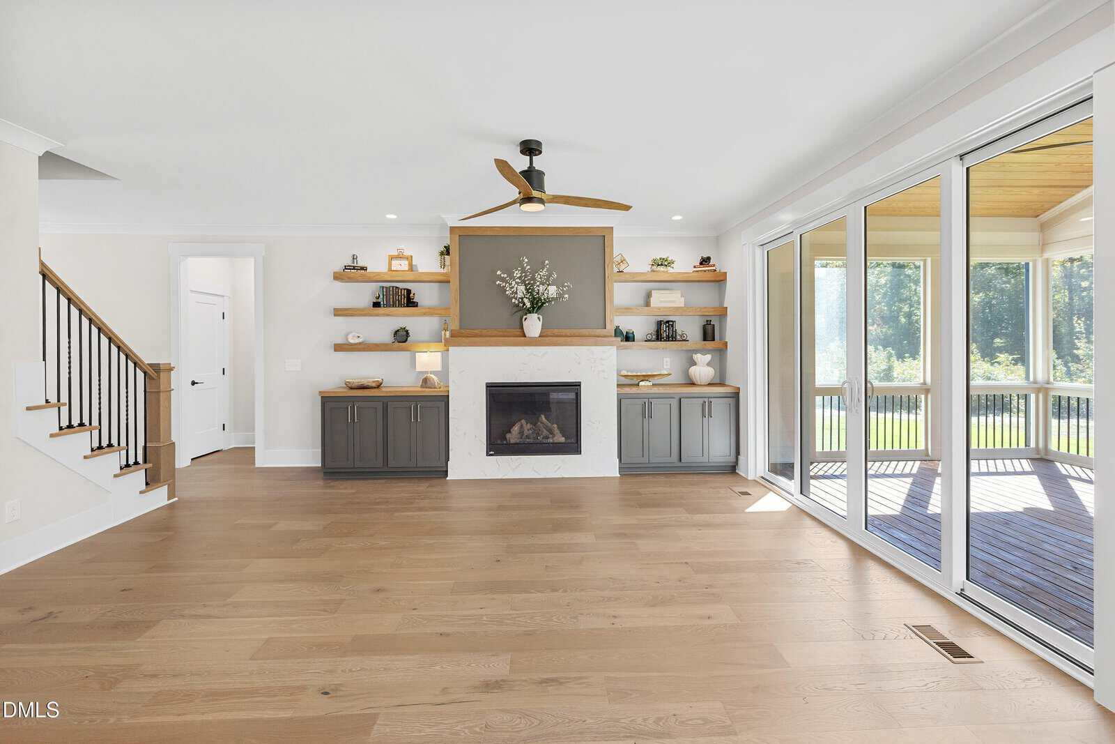 Bright living room with light wood floors, fireplace with mantel, built-in shelves and cabinets, ceiling fan, and large sliding glass doors opening to screened porch with view of trees. Stairs are visible on the left.