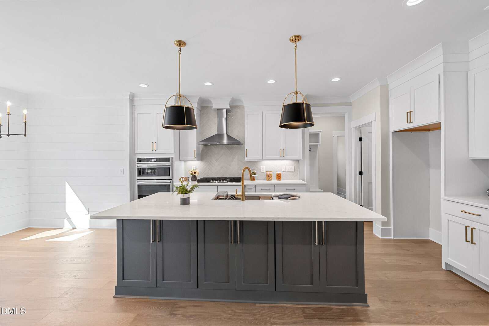 Modern kitchen with a large island featuring a white countertop, dark gray base, gold fixtures, two black pendant lights, white cabinetry, stainless steel appliances, wood flooring, and bright natural light.