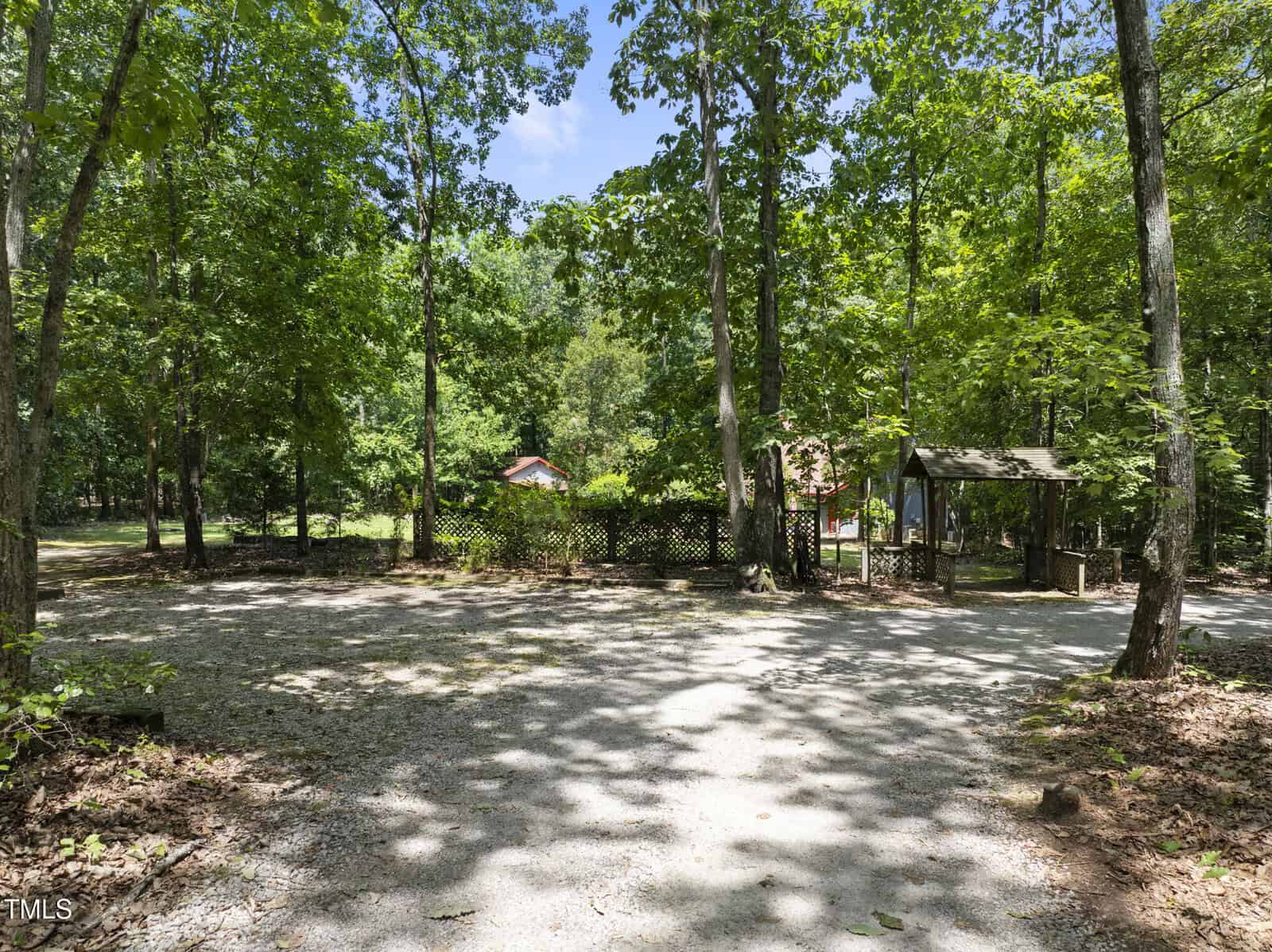 A gravel driveway surrounded by tall, green trees leads to a small wooden structure and a fenced area with a building partially visible in the background on a sunny day.