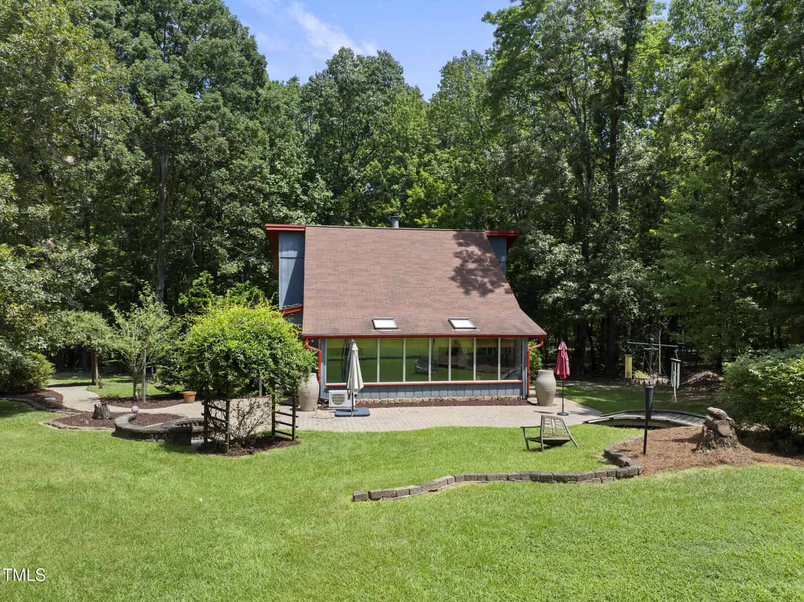 A modern house with a sloped roof sits in front of a wooded area at 4083 Graham Sherron Road. The neatly mowed lawn features landscaping, a stone patio, outdoor furniture, and a red umbrella in the foreground near Wake Forest.