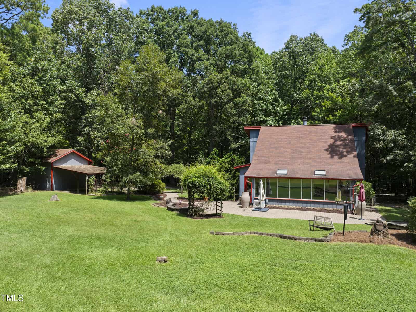 A two-story house with a brown roof and large sunroom windows sits in a grassy yard surrounded by trees. A detached carport or shed is visible to the left, with a patio area and swing in the backyard.