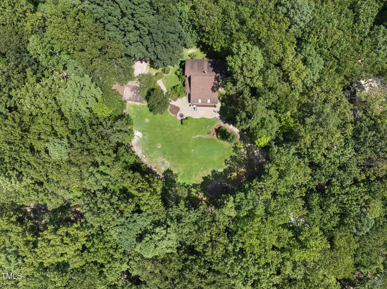 Aerial view of a house with a red roof surrounded by dense green trees and a large grassy yard, located in the center of a forested area. Paths and landscaping are visible near the house.