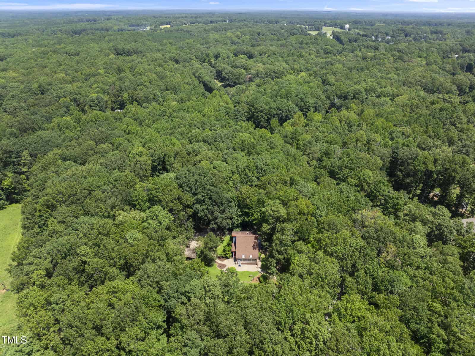 Aerial view of a house surrounded by dense, green forest. The property is mostly hidden by trees, and the landscape stretches to the horizon under a partly cloudy sky.