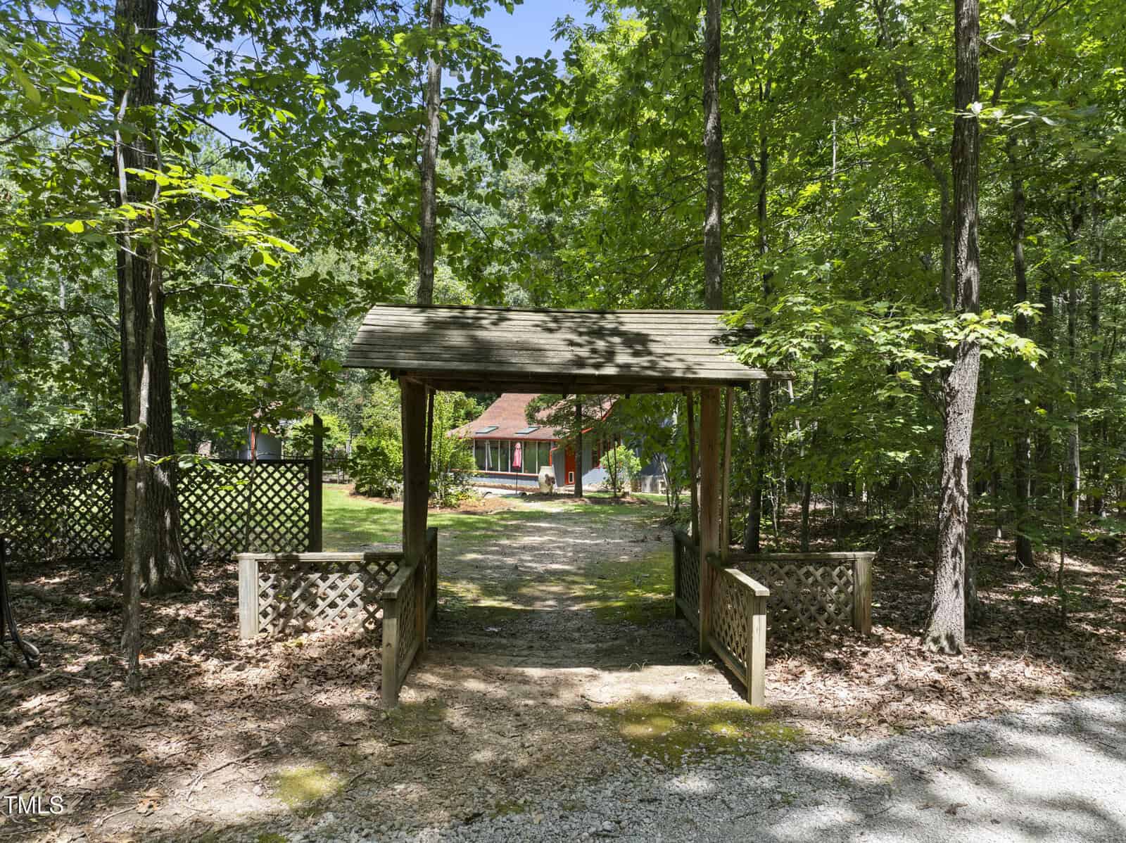 A wooden canopy structure stands over a gravel driveway leading to a house with a red roof, surrounded by green trees and shaded by forest. Lattice panels border the structure, and sunlight filters through the trees.