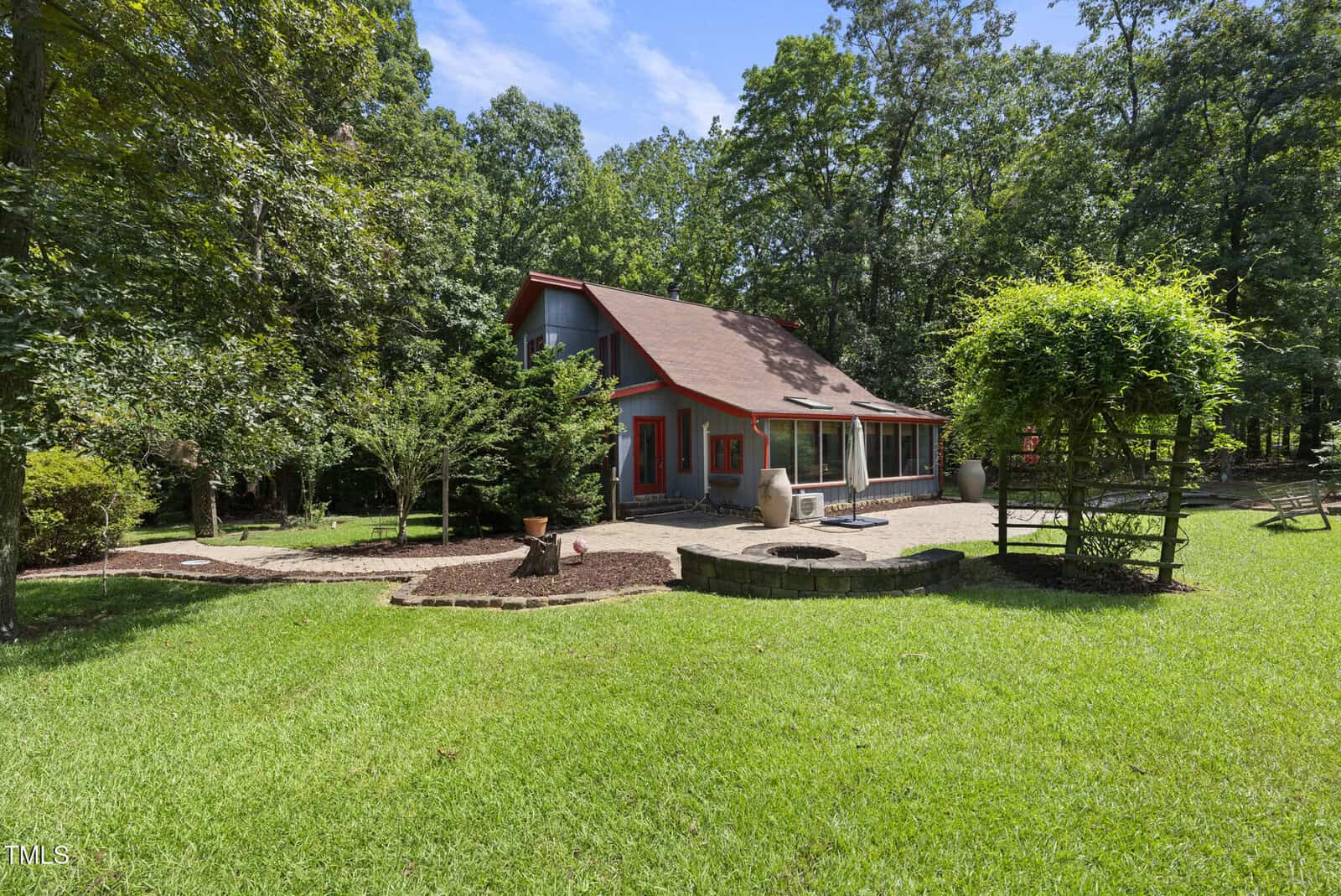 A two-story house with red trim sits in a grassy yard surrounded by tall trees. The backyard features a large patio, outdoor furniture, a fire pit, and a wooden pergola covered with greenery.