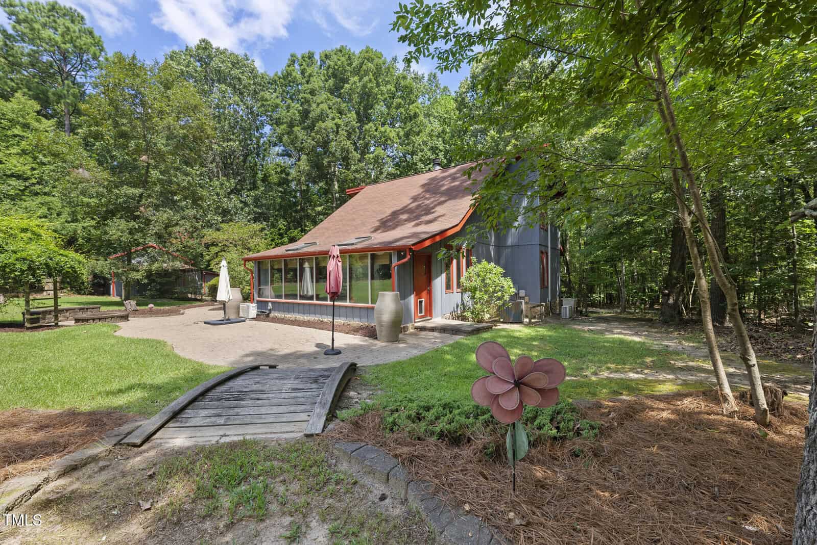 A red and gray two-story house with a sloped roof is surrounded by trees. There is a small wooden footbridge, an umbrella patio set, and a large pink metal flower sculpture in the landscaped yard.