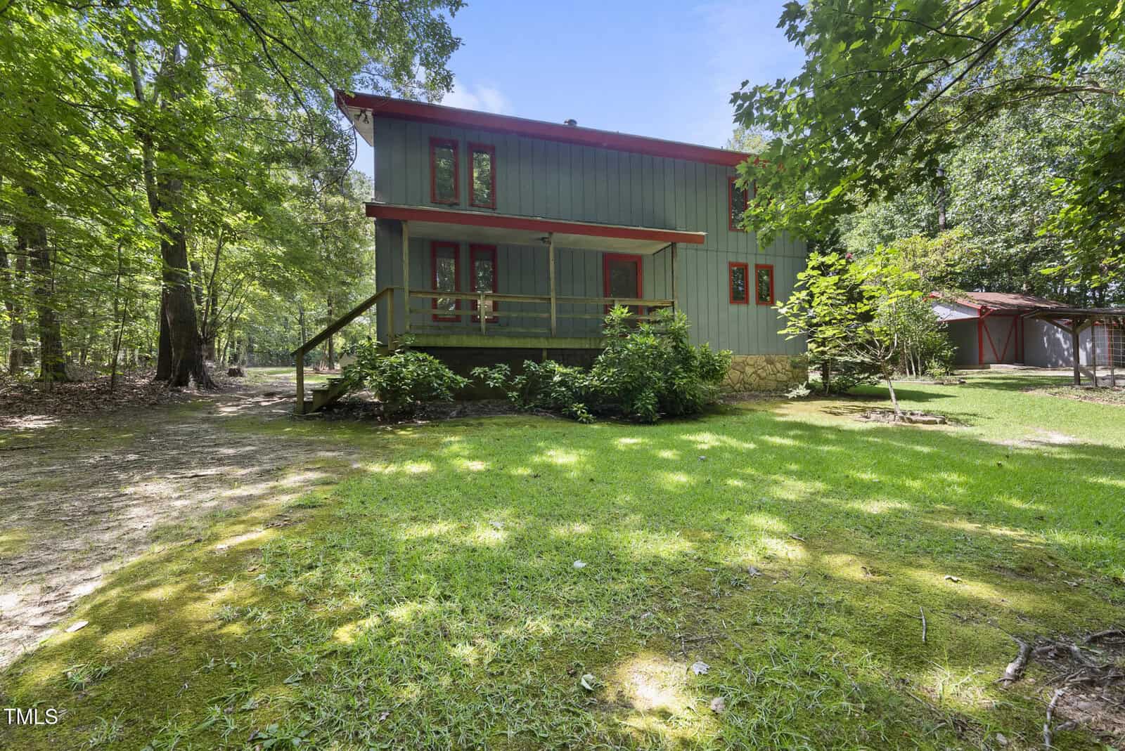 A two-story house with green vertical siding and red trim sits in a wooded area. The yard has green grass and mature trees, and a wooden shed is visible in the background to the right.