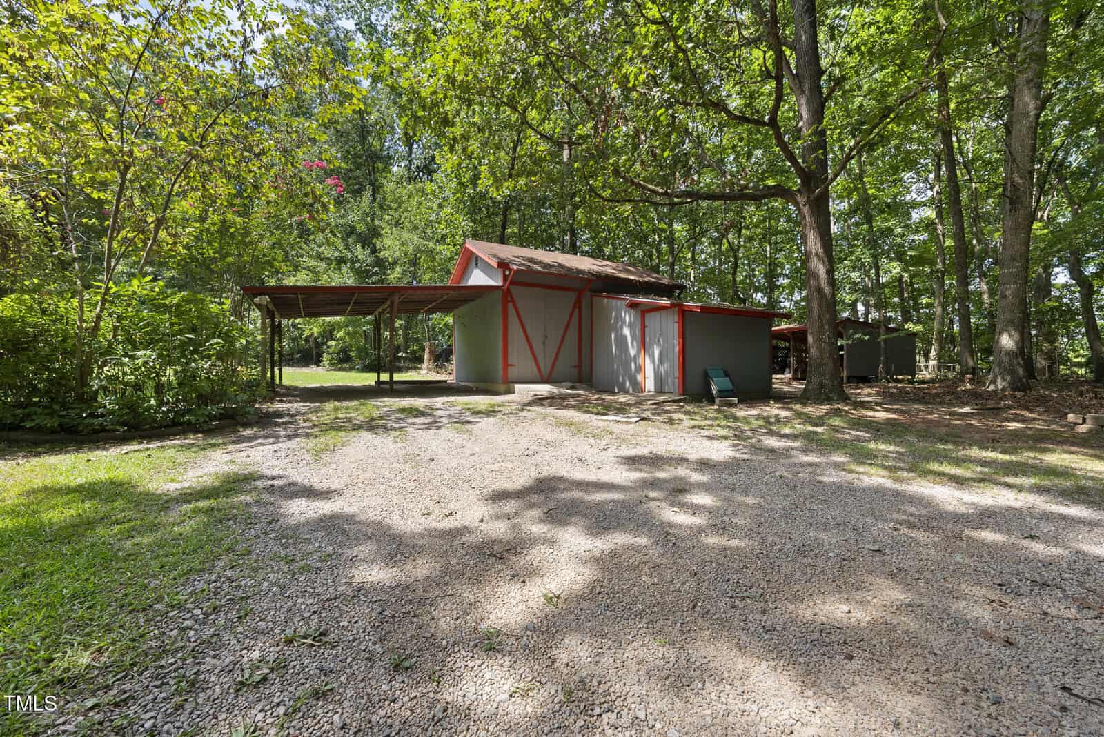 A gray and red barn-style shed sits in a wooded area with a gravel driveway. To the left is a covered carport. Shadows from tall trees cover the ground, and green grass borders the gravel driveway.