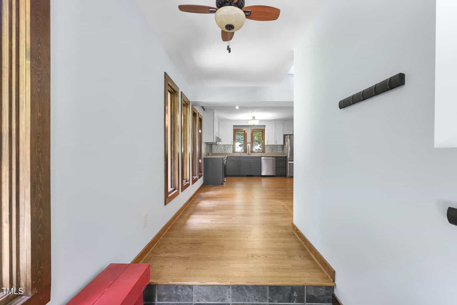 A hallway with wooden floors leads to a modern kitchen. Large windows line the left wall, and a ceiling fan hangs above. A red bench sits on the left, and a coat rack is on the right wall.