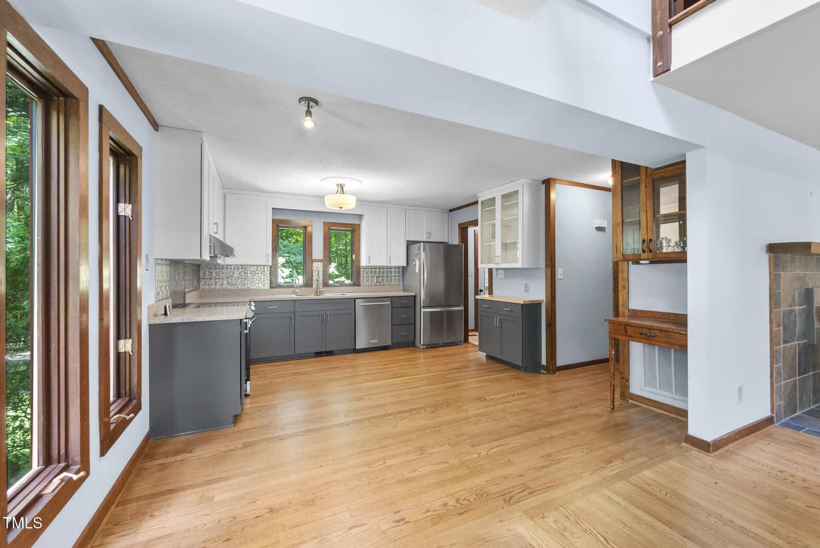 Modern kitchen with gray lower cabinets, white upper cabinets, stainless steel appliances, light wood floor, and large windows letting in natural light. Adjacent dining area and partial view of a fireplace.