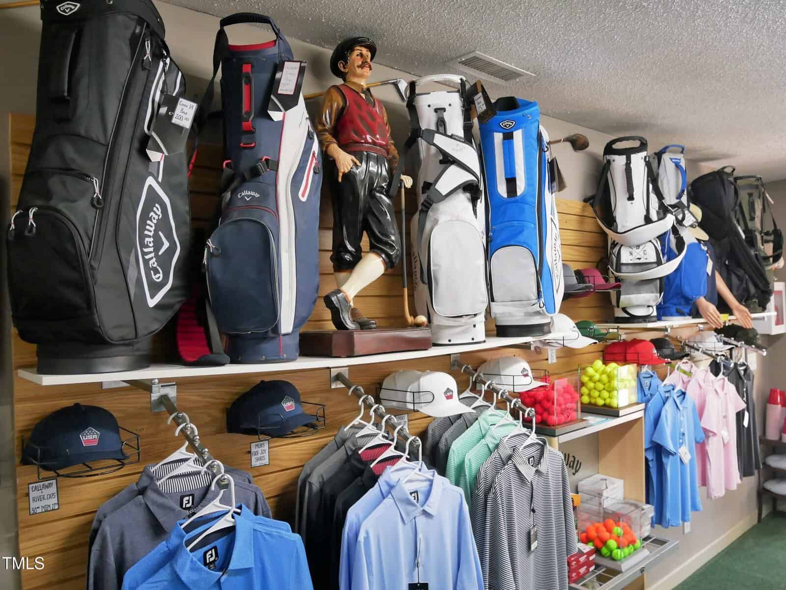 Golf bags, hats, shirts, and other accessories are neatly displayed on shelves and racks in a golf pro shop. A wooden golfer figurine stands among the bags. Golf balls and folded clothing are also visible.