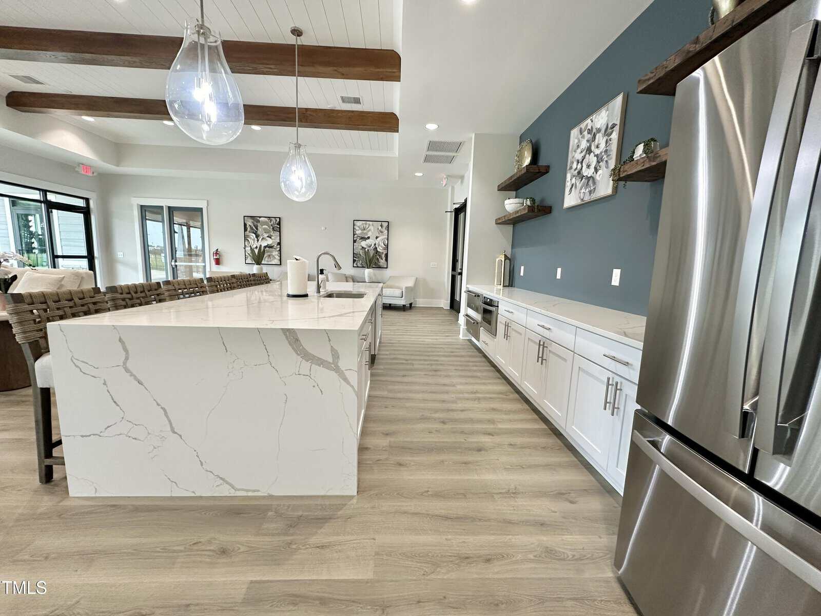 Modern kitchen with white marble island, stainless steel appliances, white cabinets, wood flooring, teal accent wall, floating shelves, two pendant lights, and exposed wood ceiling beams. Large windows let in natural light.