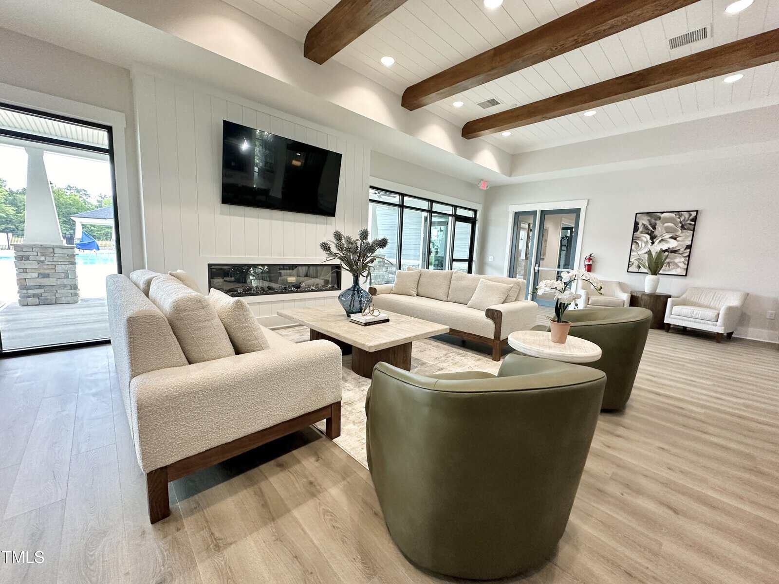 Modern living room with beige sofas, green armchairs, a coffee table, large TV, fireplace, wooden ceiling beams, and floor-to-ceiling windows overlooking a pool area. Decor includes vases with plants.