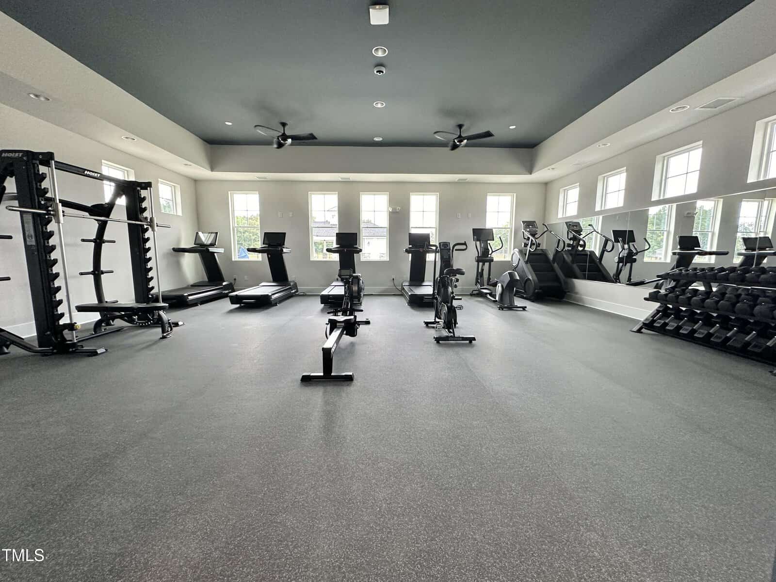A spacious gym room with weight racks, treadmills, rowing machines, stair climbers, and large windows along two walls letting in natural light. The floor is covered in rubber matting.
