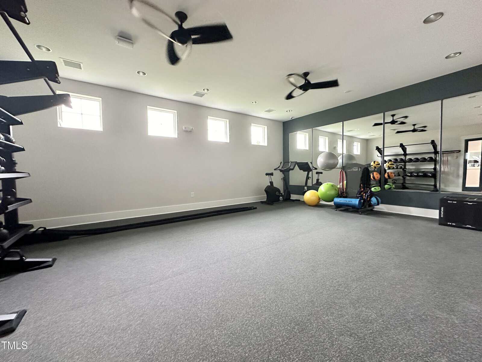 A home gym with gray flooring, mirrored wall, exercise equipment including weights, stability balls, mats, and stationary bikes. There are three ceiling fans and four small windows on one wall.