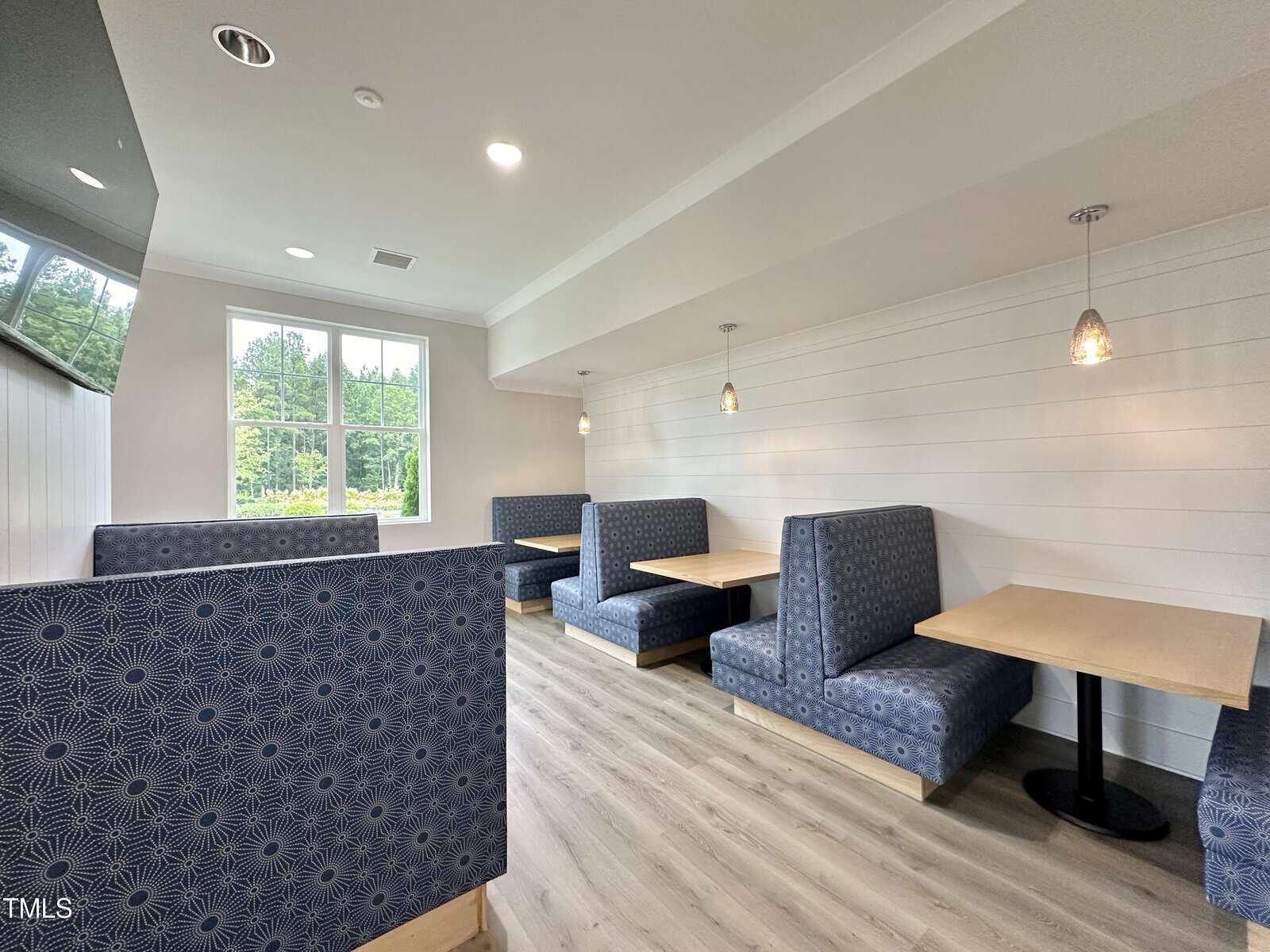 A dining area with four blue upholstered booth seats and wooden tables, pendant lights above each booth, light wood flooring, white walls, and a large window providing natural light.