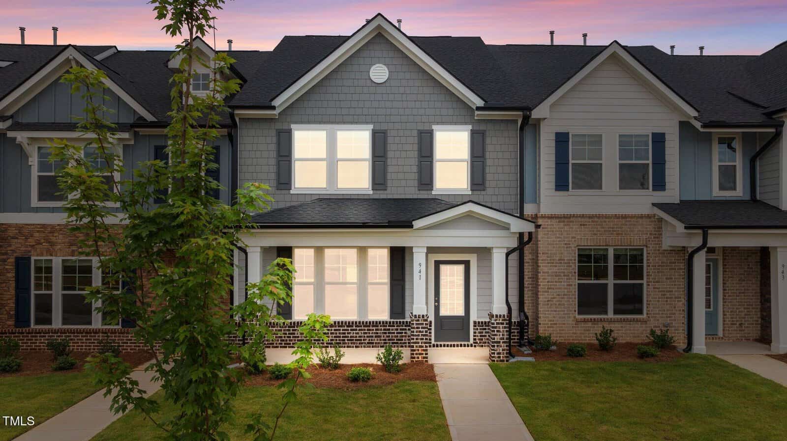 A modern two-story townhouse with gray siding, brick accents, white trim, and large windows at sunset. A small front porch and walkway lead to the entrance, with landscaped shrubs and a young tree in the front yard.