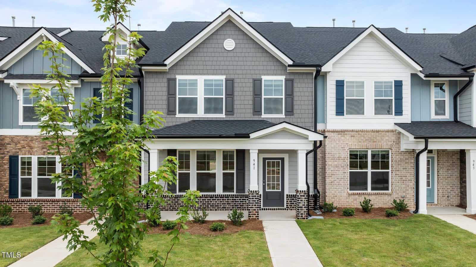 A modern two-story townhouse with gray siding, white trim, blue shutters, a covered front porch, and a small, landscaped front yard with a young tree and neatly trimmed grass.