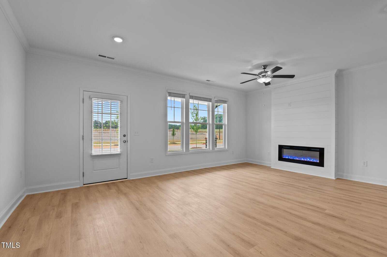 Empty living room with light wood flooring, white walls, three large windows, a glass door, ceiling fan with lights, and a modern electric fireplace built into the wall.