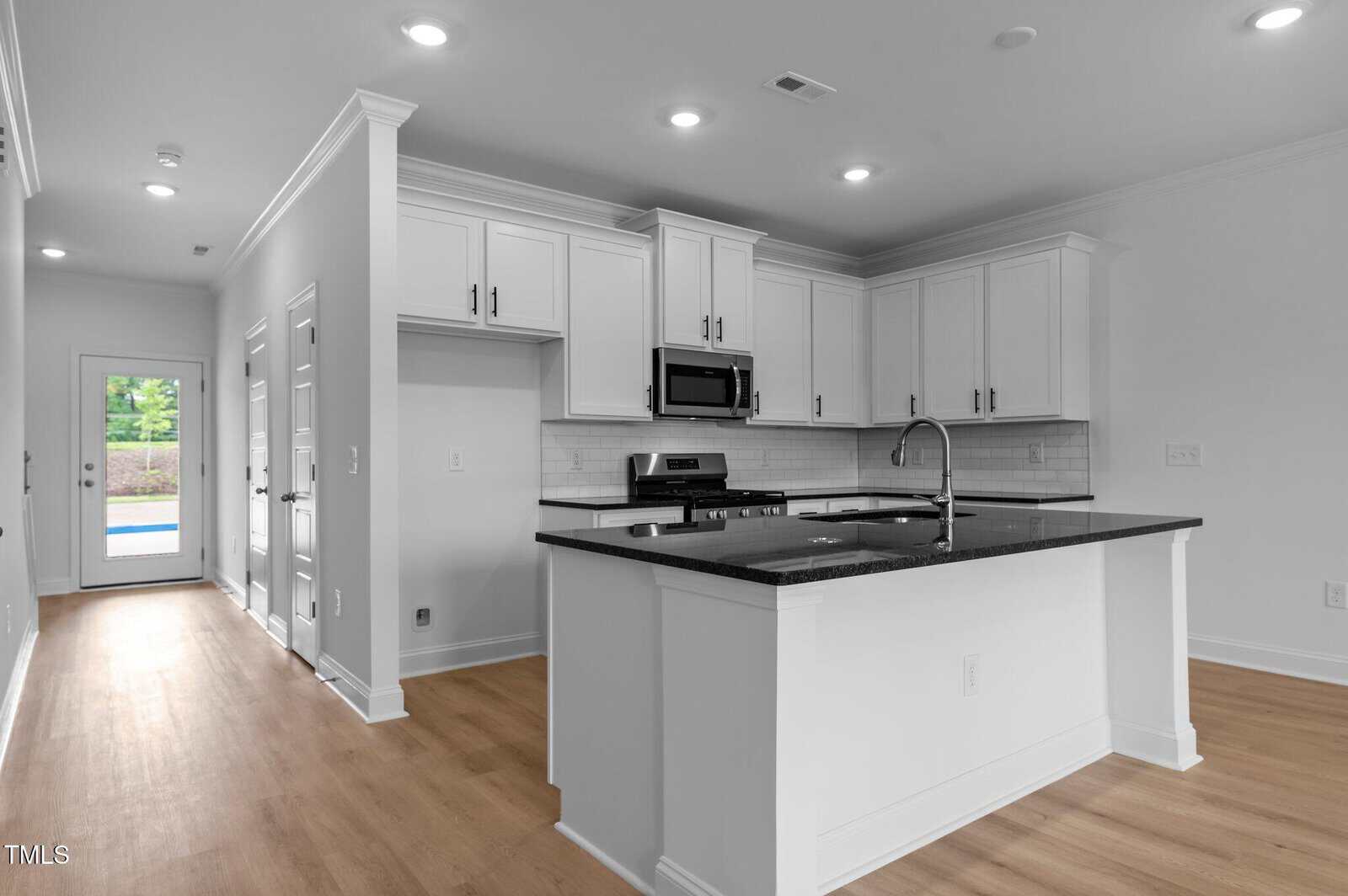 Modern kitchen with white cabinets, black countertops, stainless steel appliances, a center island with a sink, and light wood flooring. A hallway leads to a glass door with a view of greenery outside.