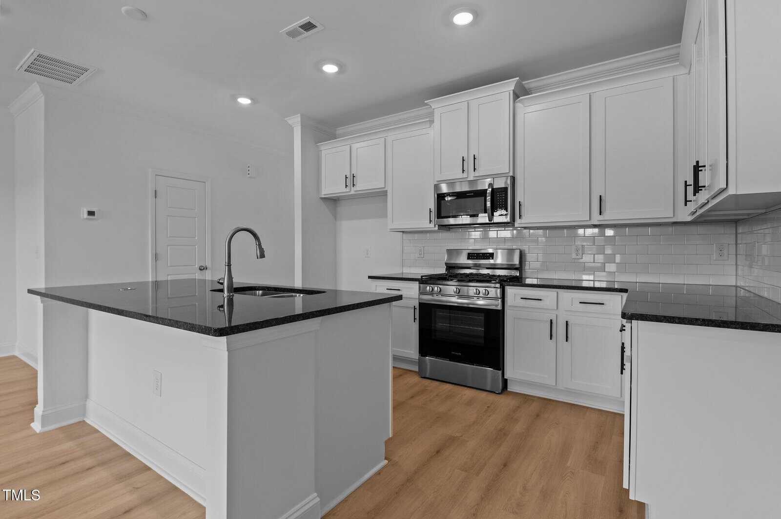A modern kitchen with white cabinets, black countertops, a central island with a sink, stainless steel appliances, a subway tile backsplash, and light wood flooring.