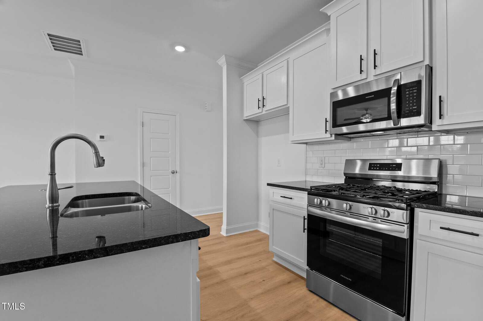 Modern kitchen with white cabinets, black granite countertops, stainless steel appliances including a gas stove and microwave, subway tile backsplash, and light wood flooring. An island with a sink is in the foreground.