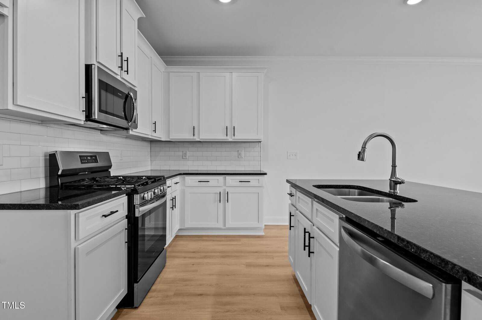 Modern kitchen with white cabinets, stainless steel appliances, a gas stove, black countertops, a tile backsplash, and a central island with a sink and dishwasher. The floor is light wood, and the lighting is recessed.