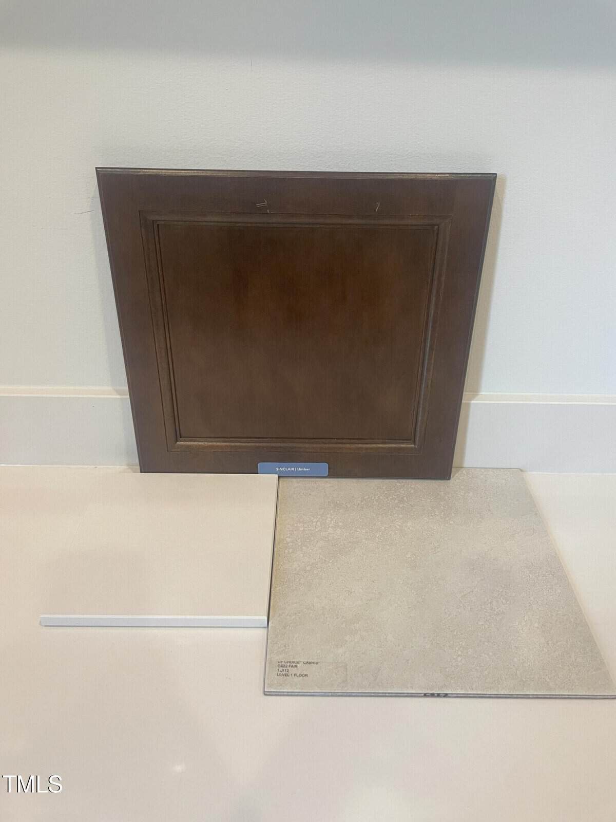 A dark brown cabinet door stands upright against a wall, with a beige square floor tile and a thin white strip arranged on a light-colored surface in front of it.
