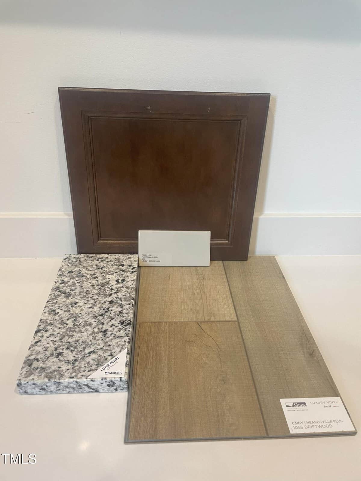 A brown wood cabinet door, a light gray paint swatch, a granite countertop sample with black and white speckles, and a wood-look vinyl flooring sample are displayed upright against a light wall.
