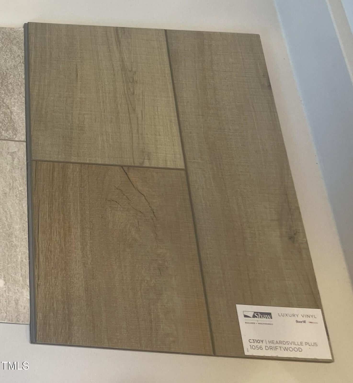 Sample display of luxury vinyl flooring in a wood-look pattern, featuring neutral brown and gray tones with visible grain lines. A label in the corner reads “Luxury Vinyl, 1056 Driftwood.”.