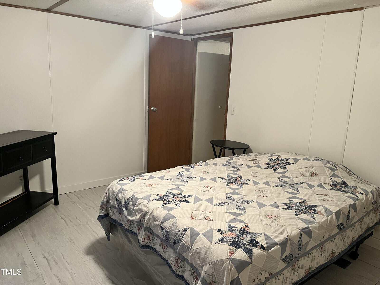A simple bedroom with white walls, a ceiling fan, a wooden door, a bed covered with a patchwork quilt, a small round side table, and a black desk with shelves. The floor appears to be light-colored tile or laminate.
