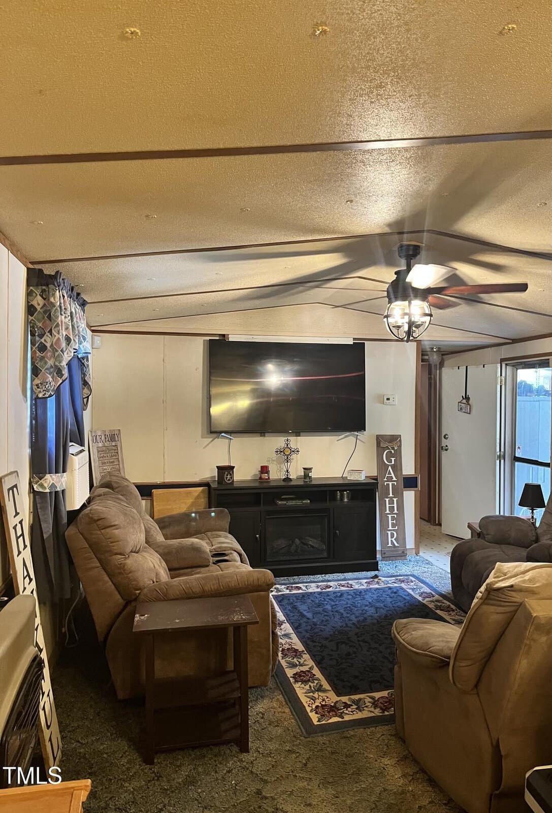 Living room with beige walls, tan reclining sofas, a wood side table, a fireplace with decorations, a flat-screen TV, a ceiling fan with lights, a blue area rug, and a window with patterned curtains. Door visible in the background.