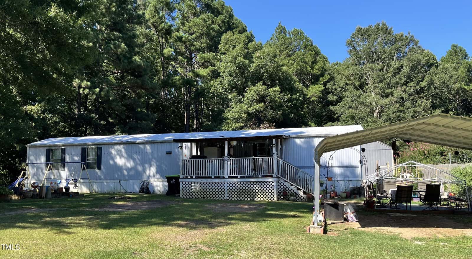 A long white mobile home with a covered porch sits on a grassy yard surrounded by trees. To the right, there is a metal carport sheltering a patio table and chairs. Bright sunlight casts shadows across the scene.