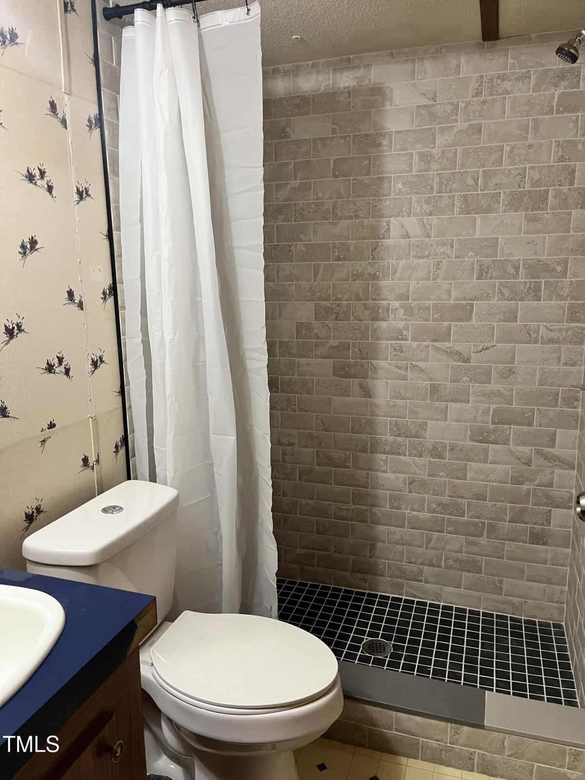 A bathroom with a white toilet, a sink with a blue countertop, a shower with a white curtain, and tan tile walls. The shower floor is black tile. The wallpaper has a floral pattern.