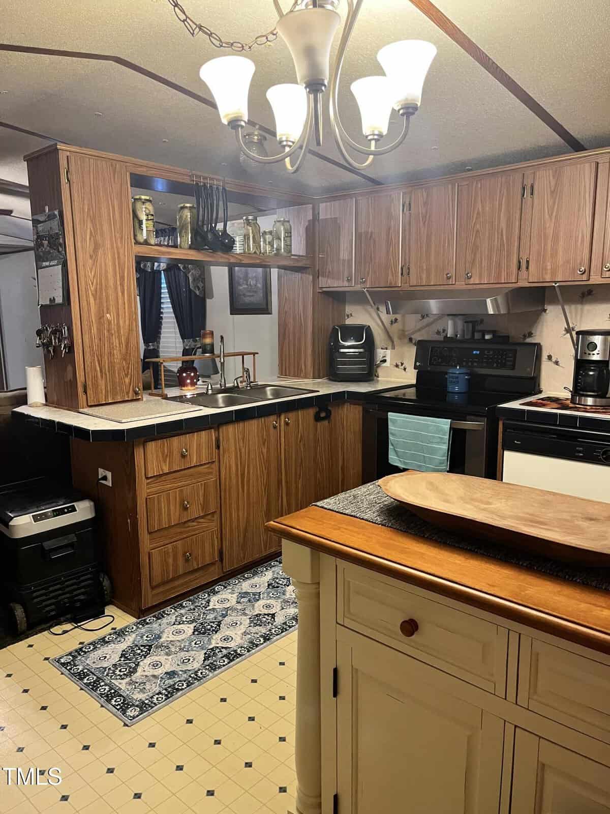 A cozy kitchen with wood cabinets, a stove, microwave, coffee maker, and dish rack. A blue patterned rug lies on the floor, and a cream-colored island with a wooden tray sits in the foreground under a chandelier.