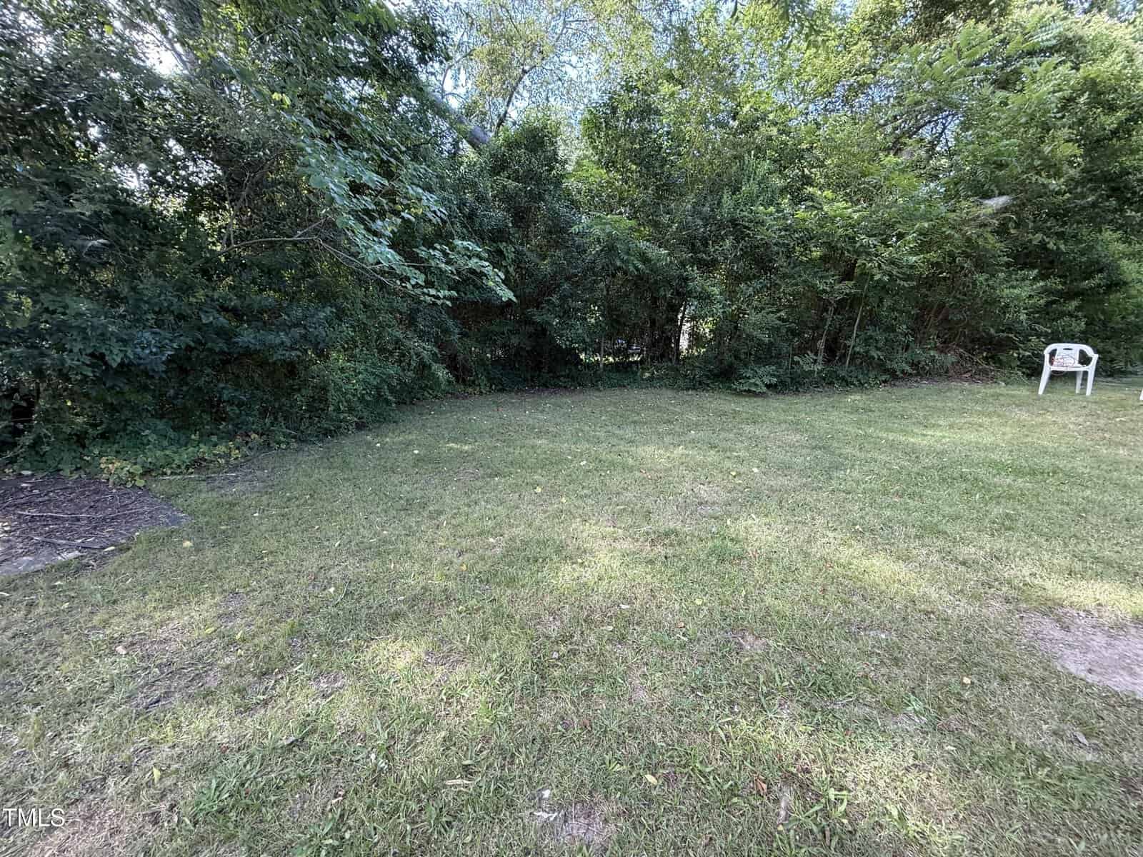 A grassy backyard with a patch of dirt on the left, dense green trees and bushes in the background, and a single white plastic chair near the right edge of the image.