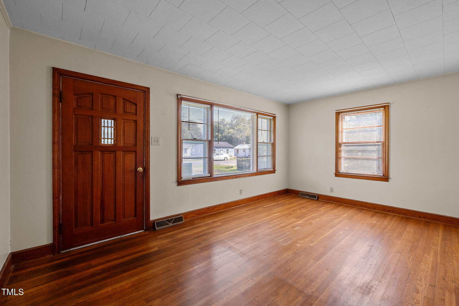 Unfurnished room with hardwood floors, a wooden front door with glass panes, beige walls, a white tile ceiling, and three windows letting in natural light. Exterior view through windows shows houses and vehicles.