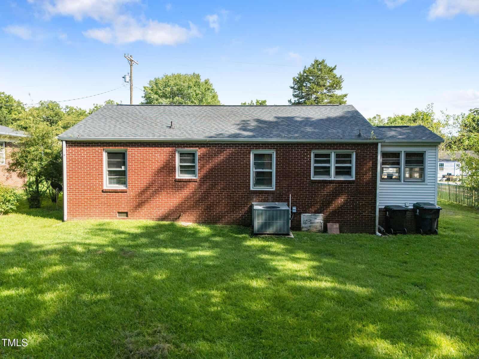 A red brick single-story house with a gray roof, three windows, an HVAC unit, and a grassy backyard under a blue sky with some clouds.