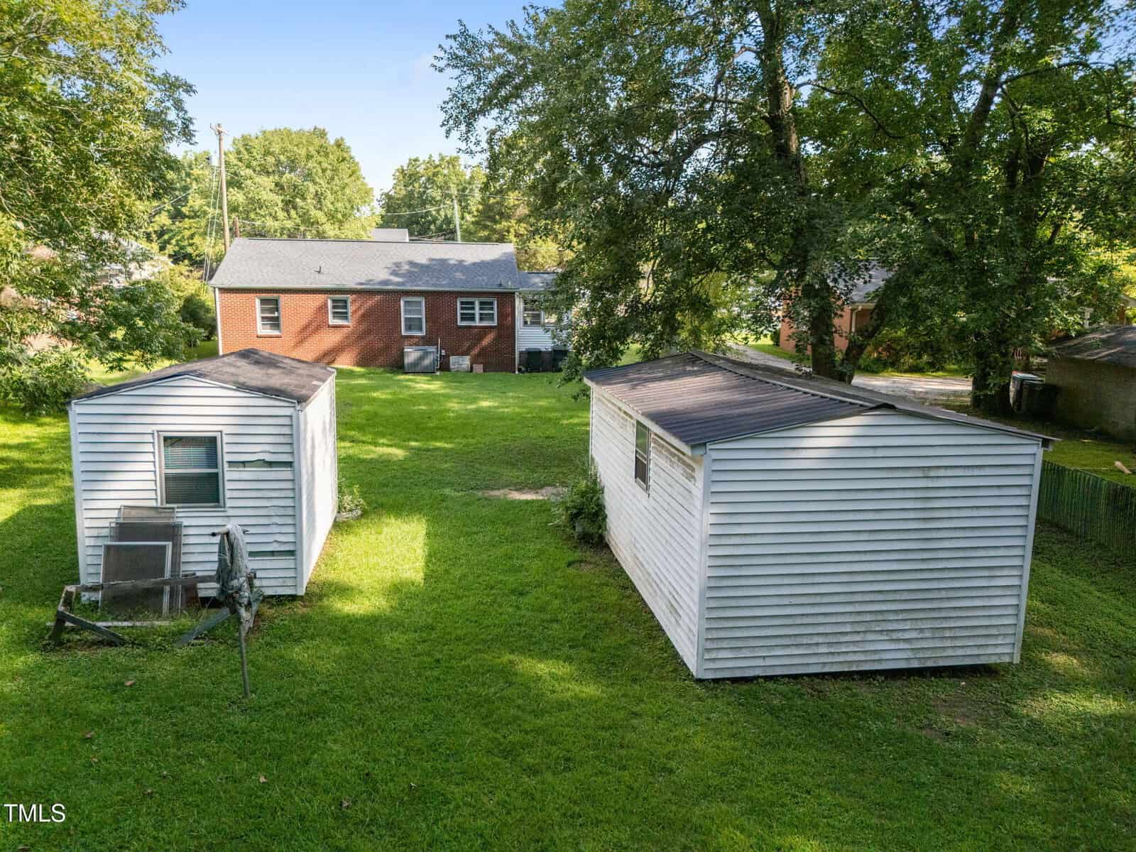 A backyard with green grass, two white sheds, and a brick house in the background, surrounded by trees on a sunny day.