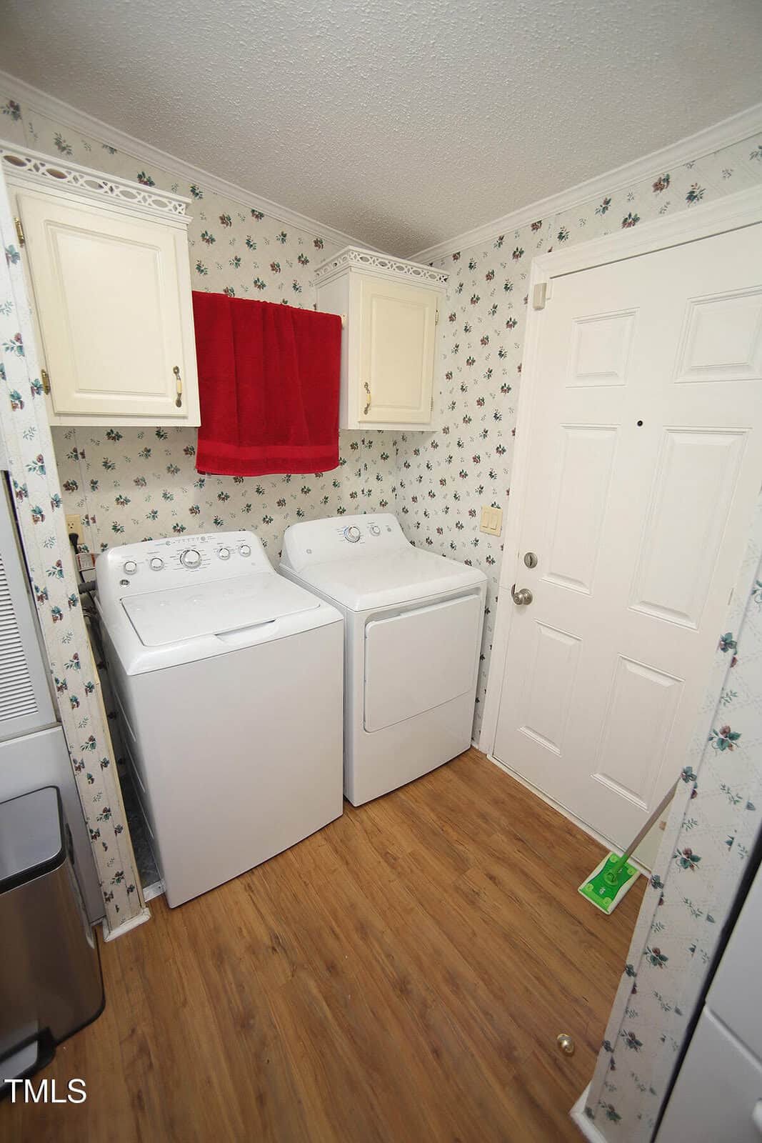 A laundry room with a white washing machine and dryer side by side, wooden floor, white cabinets, a red towel hanging between the cabinets, floral wallpaper, and a white door with a broom nearby.